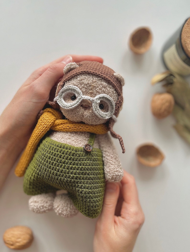 May include: A crocheted plush toy bear wearing a brown aviator hat, round glasses, a yellow scarf, and green overalls with a brown button.
