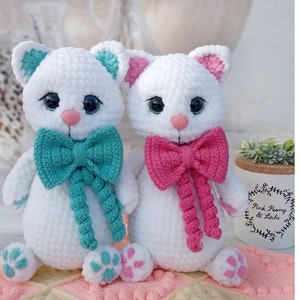 May include: Two crocheted white cat plushies with embroidered eyes and pink and turquoise bows. The plushies are sitting on a pink and white striped blanket.