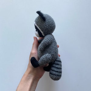 May include: A gray crocheted raccoon plush toy with black eyes and a black and gray striped tail. The toy is being held in a person's hand.