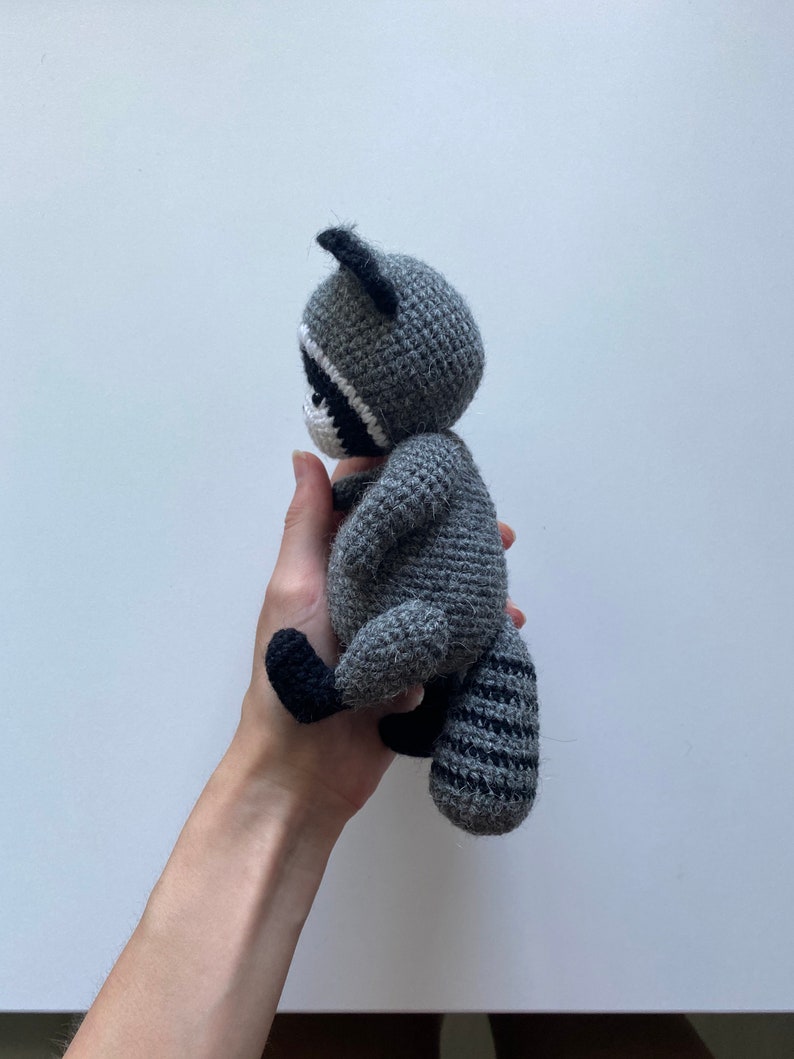 May include: A gray crocheted raccoon plush toy with black eyes and a black and gray striped tail. The toy is being held in a person's hand.
