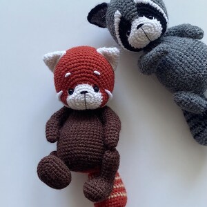 May include: Two crocheted stuffed animals, a red panda and a raccoon. The red panda is brown with a red head and a striped tail. The raccoon is gray with a black mask.