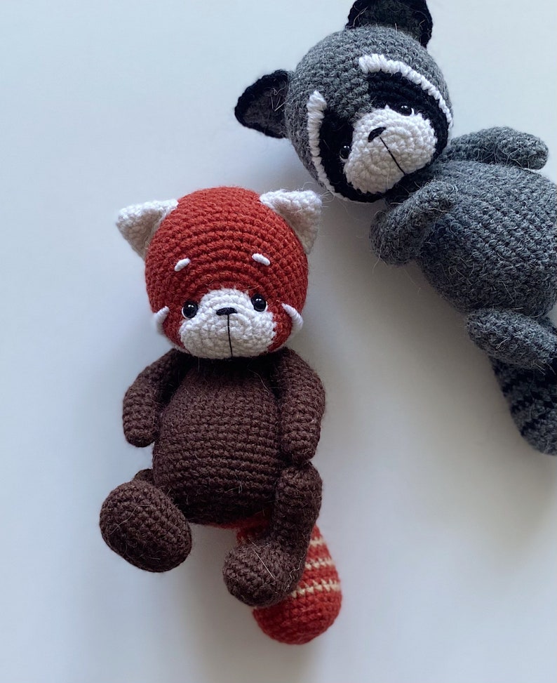 May include: Two crocheted stuffed animals, a red panda and a raccoon. The red panda is brown with a red head and a striped tail. The raccoon is gray with a black mask.