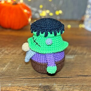 May include: A crocheted Frankenstein monster toy with green skin, black hair, and purple clothing. The toy has a stitched scar on its face and is holding a small white crocheted object.