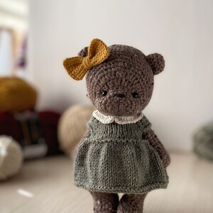 May include: A brown crocheted teddy bear wearing a green dress with a white collar and a yellow bow. The bear is standing on a light wood surface.