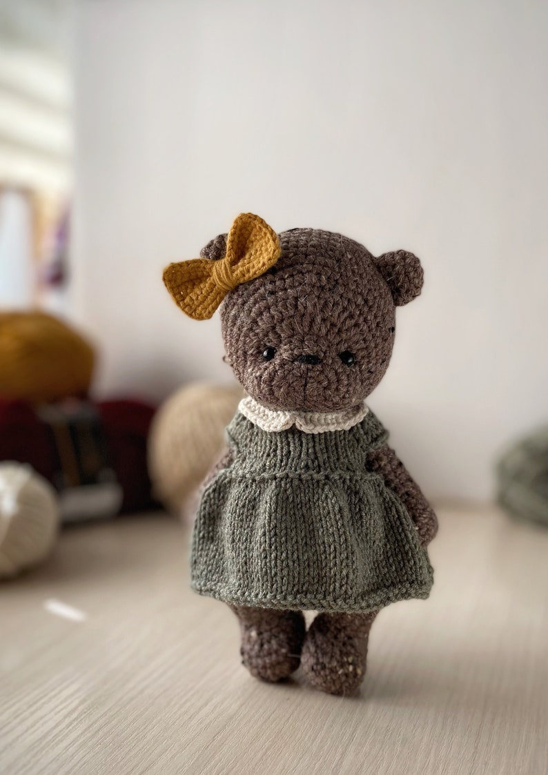 May include: A brown crocheted teddy bear wearing a green dress with a white collar and a yellow bow. The bear is standing on a light wood surface.