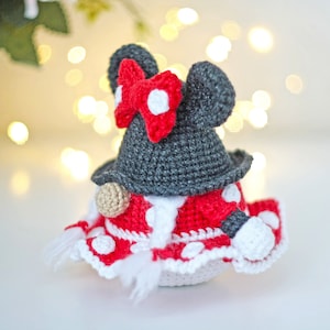 May include: A crocheted gnome doll with a red and white polka dot dress, a black hat with white polka dots, and a red bow. The gnome is standing on a white surface.