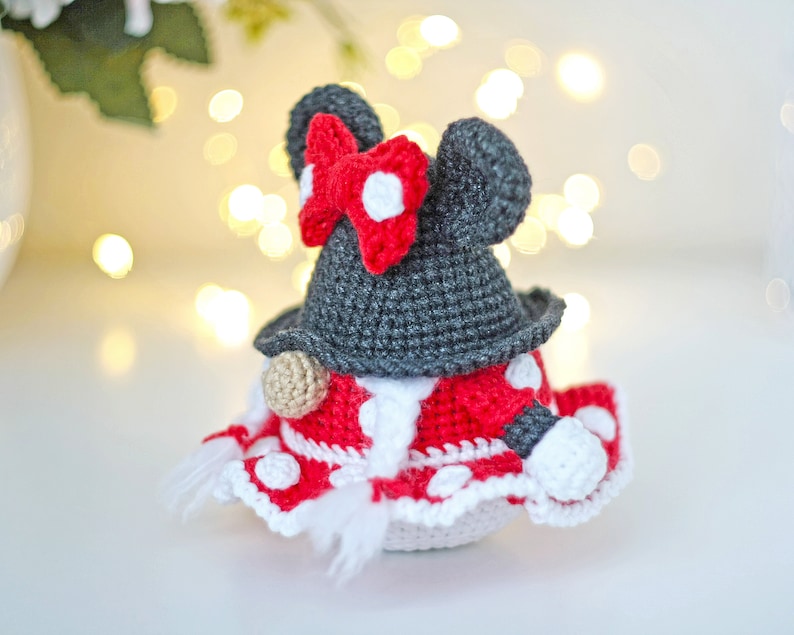 May include: A crocheted gnome doll with a red and white polka dot dress, a black hat with white polka dots, and a red bow. The gnome is standing on a white surface.