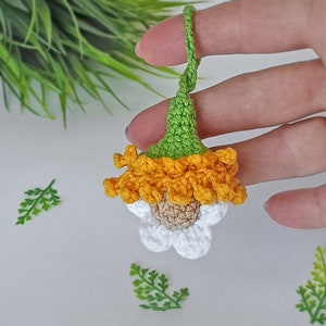 May include: A crocheted flower charm with a green stem, orange petals, and a white center. The flower is hanging from a string.
