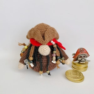 May include: Crocheted brown pirate doll with red bandana, black yarn hair, and white beads. The doll is holding a gold coin and is sitting on a white surface.