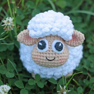 Pu&ograve; includere: Un giocattolo di pecora in crochet bianco con occhi neri e viso marrone. La pecora &egrave; seduta in un campo di erba verde.