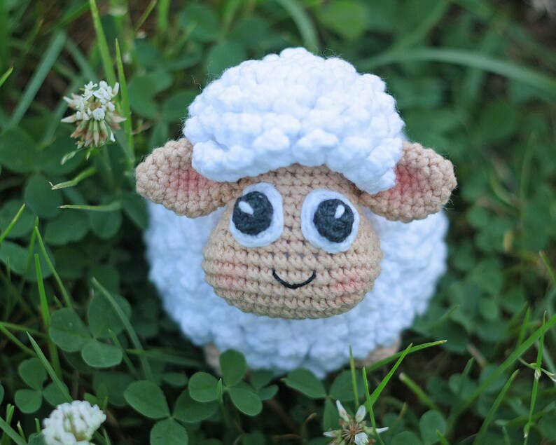 Pu&ograve; includere: Un giocattolo di pecora in crochet bianco con occhi neri e viso marrone. La pecora &egrave; seduta in un campo di erba verde.
