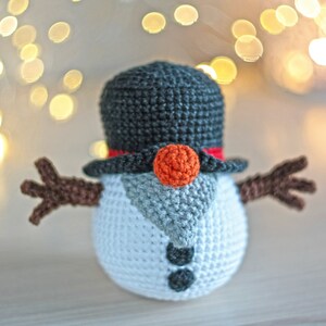 May include: A crocheted snowman with a black top hat, red trim, and an orange nose. The snowman has brown arms and a white body with gray accents.
