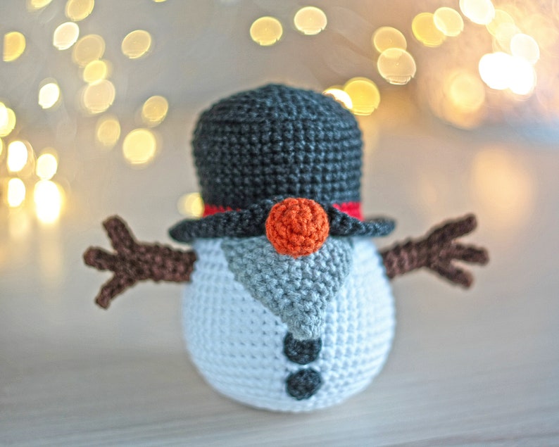 May include: A crocheted snowman with a black top hat, red trim, and an orange nose. The snowman has brown arms and a white body with gray accents.