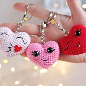 May include: Three crocheted heart-shaped keychains with smiling faces. One heart is pink with black eyes and a red mouth, another is pink with black eyes and a black mouth, and the last is red with black eyes and a black mouth.