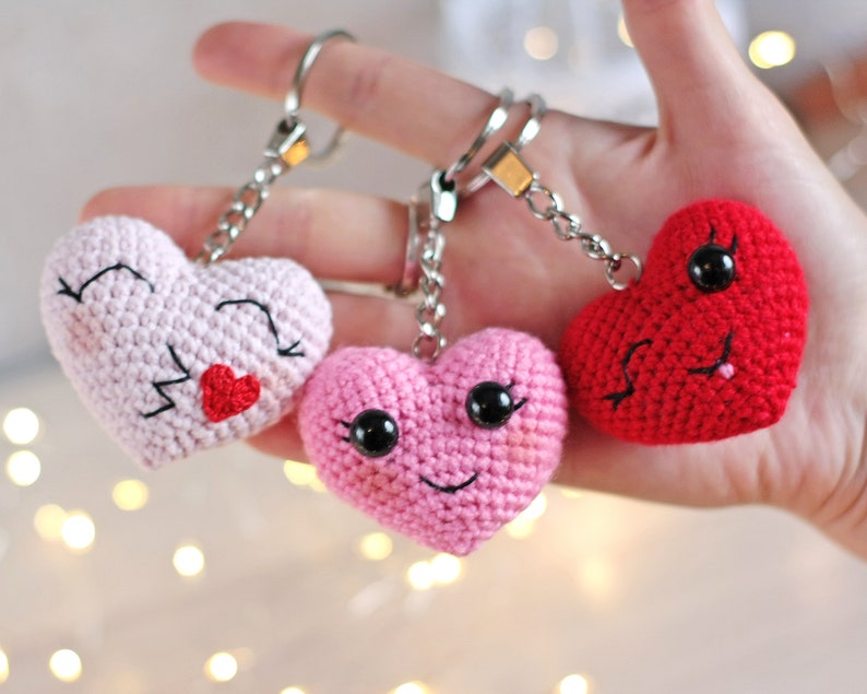 May include: Three crocheted heart-shaped keychains with smiling faces. One heart is pink with black eyes and a red mouth, another is pink with black eyes and a black mouth, and the last is red with black eyes and a black mouth.