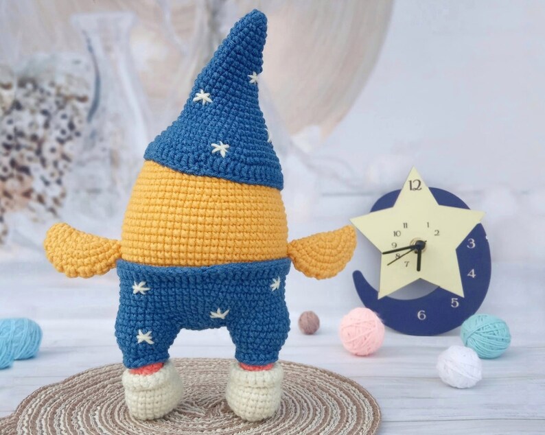 May include: A crocheted stuffed toy shaped like a person wearing a blue hat with white stars and blue pants with white stars. The toy is standing on a brown surface.
