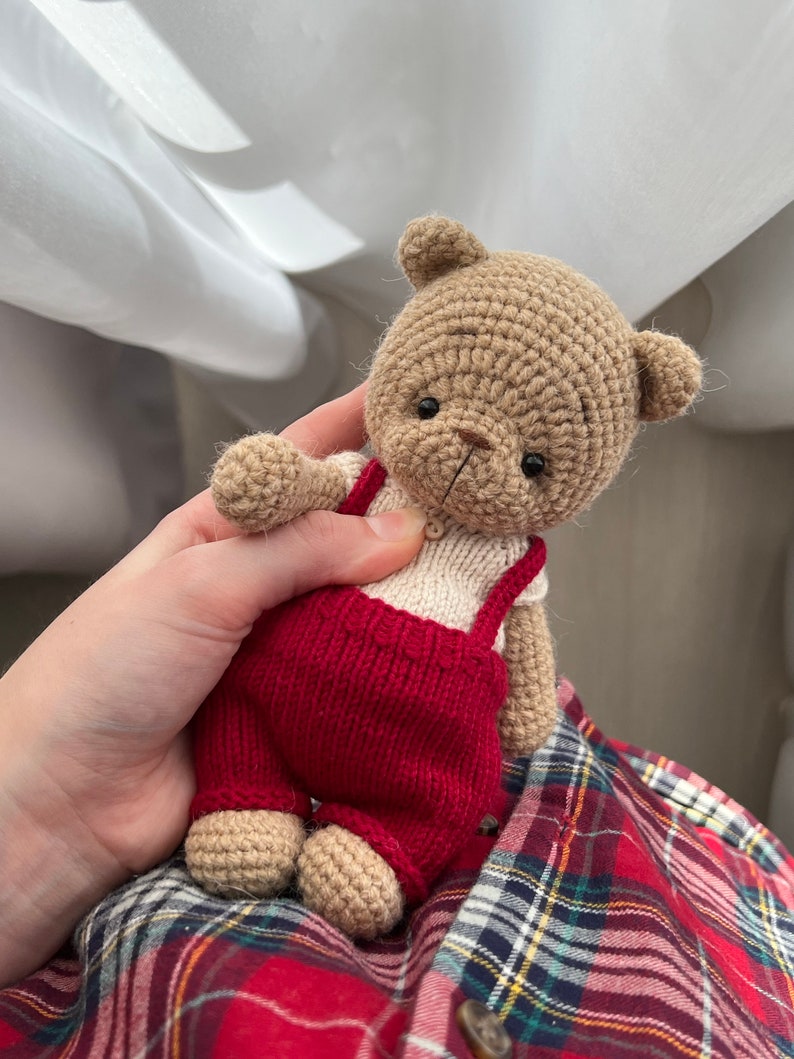 May include: A crocheted teddy bear wearing red overalls. The bear has brown fur and black eyes. The bear is being held by a hand.