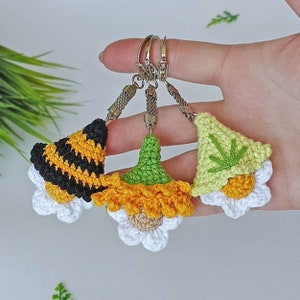 May include: Three crocheted flower keychains with different colored centers. The keychain on the left has a black and yellow center, the middle keychain has a green and orange center, and the keychain on the right has a green center.