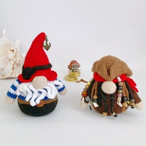 May include: Two crocheted gnomes dressed as pirates. One gnome wears a red hat with an anchor, a blue and white striped shirt, and a brown belt. The other gnome wears a brown hat with a red bandana, and has brown and gold beads around its neck.