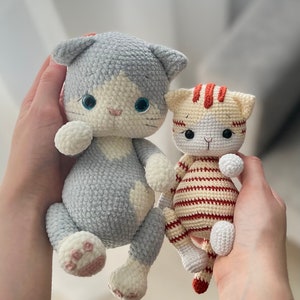 Puede incluir: Dos juguetes de gato de crochet. El gato m&aacute;s grande es gris con detalles blancos y ojos azules. El gato m&aacute;s peque&ntilde;o es naranja y blanco con rayas rojas.