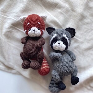 May include: Two crocheted stuffed animals, a red panda and a raccoon, are sitting on a white fabric background. The red panda has a brown body and a red head with white facial features. The raccoon has a gray body with black facial features and a black tail.
