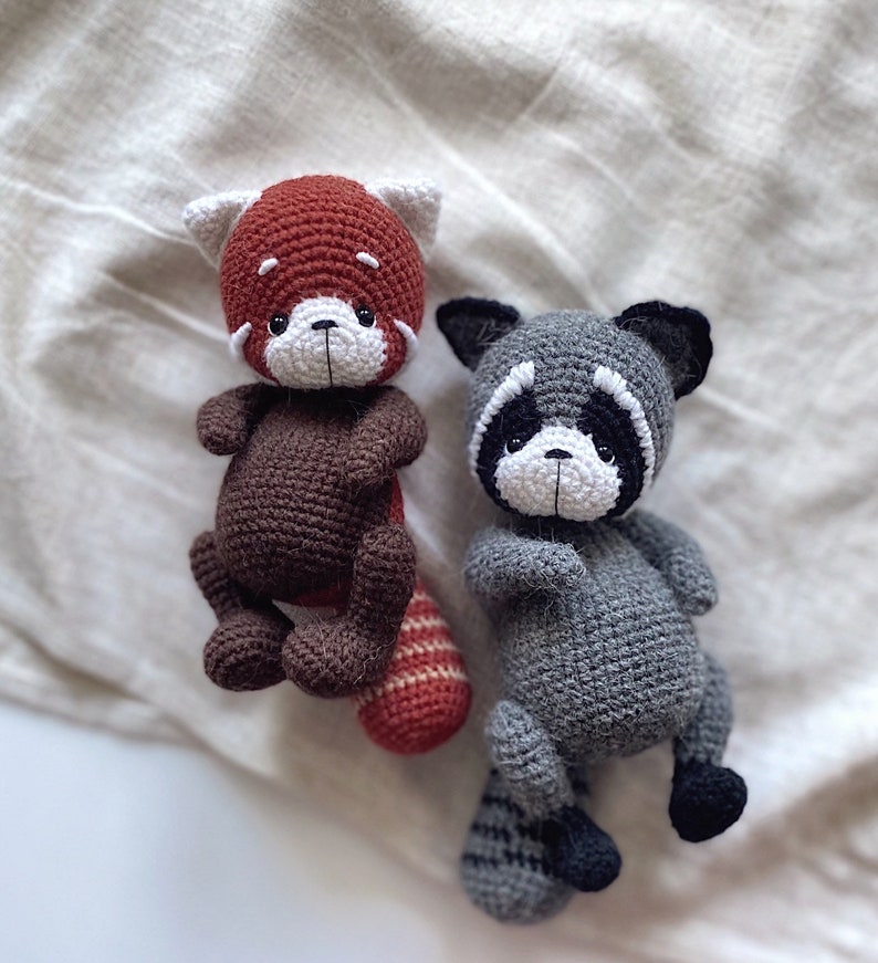 May include: Two crocheted stuffed animals, a red panda and a raccoon, are sitting on a white fabric background. The red panda has a brown body and a red head with white facial features. The raccoon has a gray body with black facial features and a black tail.