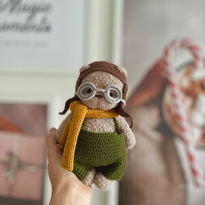 May include: A crocheted plush toy bear wearing a brown aviator hat, grey glasses, a yellow scarf, and green overalls.