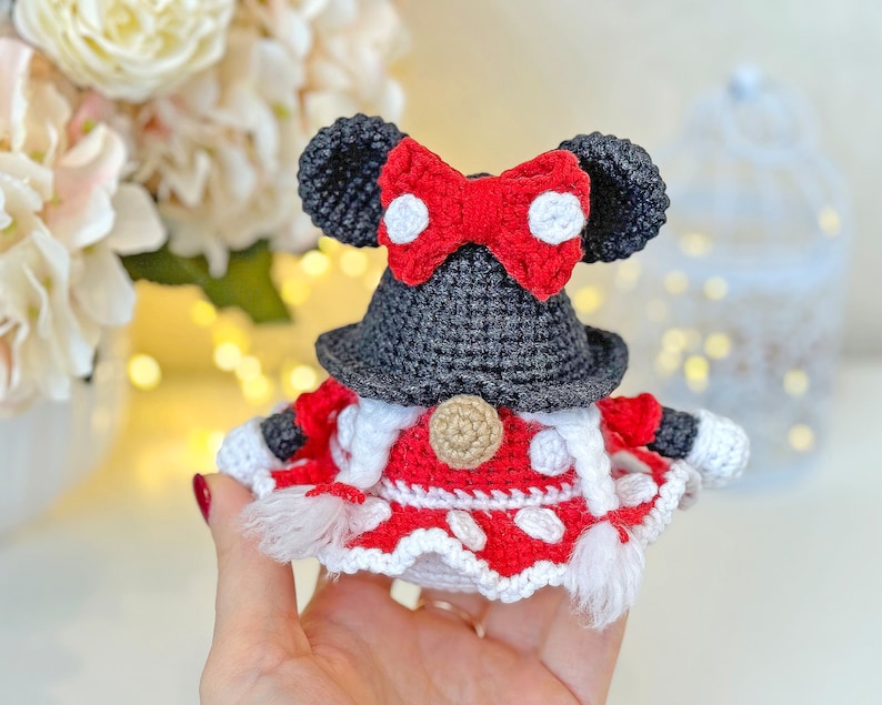 May include: A crocheted gnome doll with a red and white dress, black hat, and red bow with white polka dots. The gnome is holding a white and red striped bag.