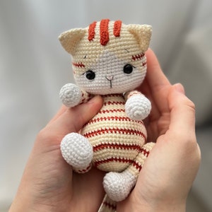 Puede incluir: Un juguete de crochet con forma de gato con rayas blancas y rojas. El gato tiene ojos negros y una cara blanca.