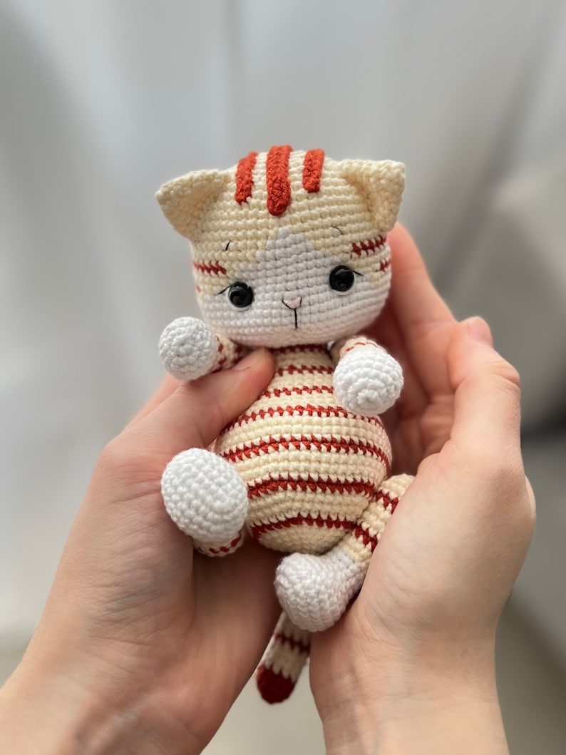 Puede incluir: Un juguete de crochet con forma de gato con rayas blancas y rojas. El gato tiene ojos negros y una cara blanca.