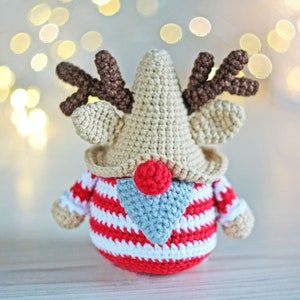 May include: A crocheted reindeer gnome with a red and white striped sweater, a brown crocheted nose, and brown antlers. The gnome is sitting on a white wooden surface.