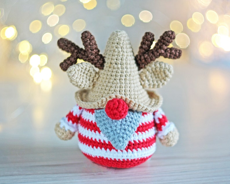 May include: A crocheted reindeer gnome with a red and white striped sweater, a brown crocheted nose, and brown antlers. The gnome is sitting on a white wooden surface.