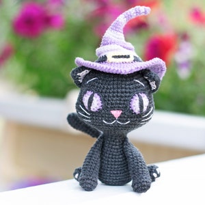 May include: A black crocheted cat wearing a purple witch hat with a black brim. The cat has white yarn for whiskers and large purple eyes.