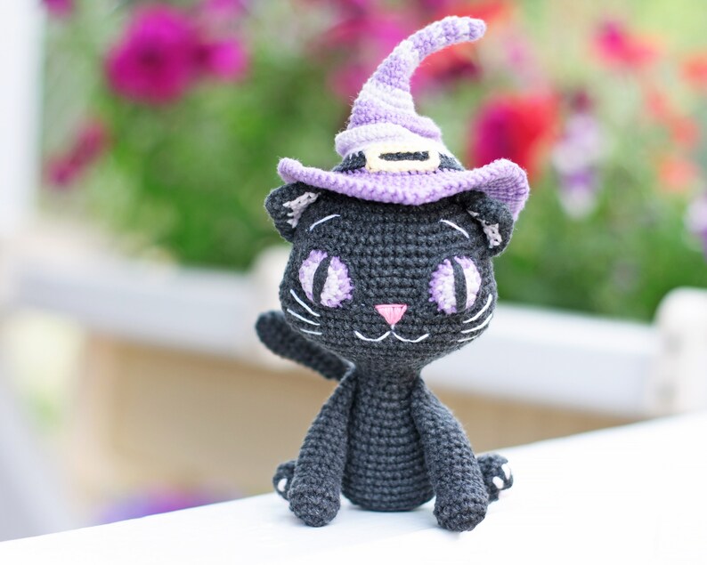 May include: A black crocheted cat wearing a purple witch hat with a black brim. The cat has white yarn for whiskers and large purple eyes.