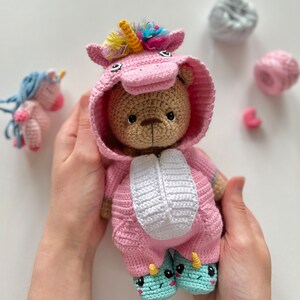 May include: A crocheted teddy bear wearing a pink unicorn costume with a rainbow horn and white accents. The bear is holding its hands together and has a sweet expression.