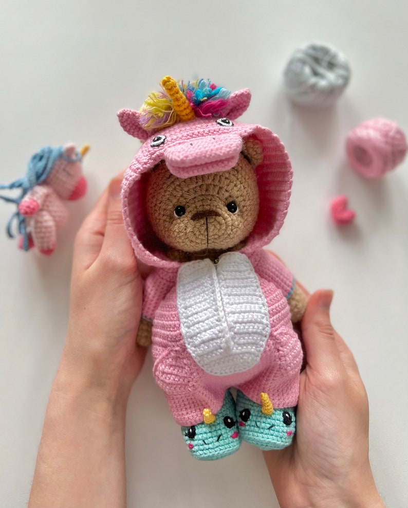 May include: A crocheted teddy bear wearing a pink unicorn costume with a rainbow horn and white accents. The bear is holding its hands together and has a sweet expression.
