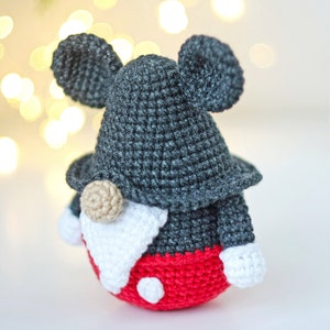 May include: A crocheted gnome doll with a gray hat and red pants. The gnome has large black ears and a white beard. The gnome is wearing a red hat with white polka dots.