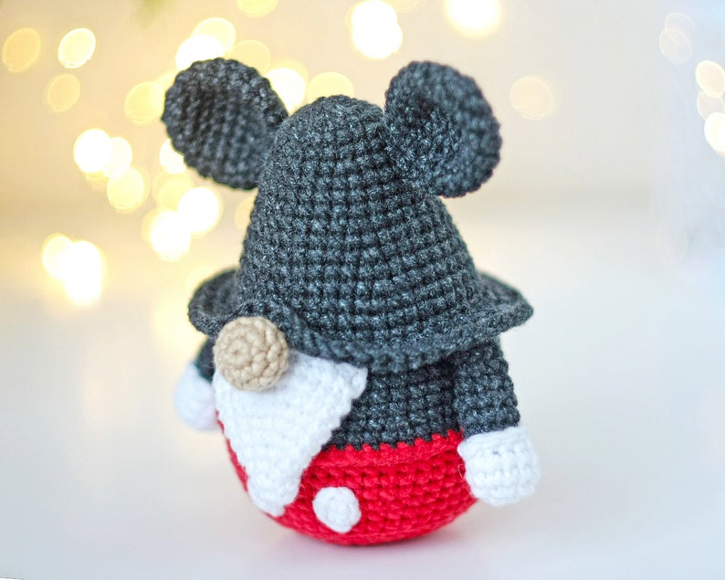 May include: A crocheted gnome doll with a gray hat and red pants. The gnome has large black ears and a white beard. The gnome is wearing a red hat with white polka dots.