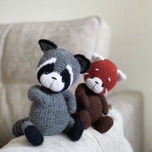 May include: Two crocheted stuffed animals, a gray raccoon and a brown red panda, sitting on a white surface.