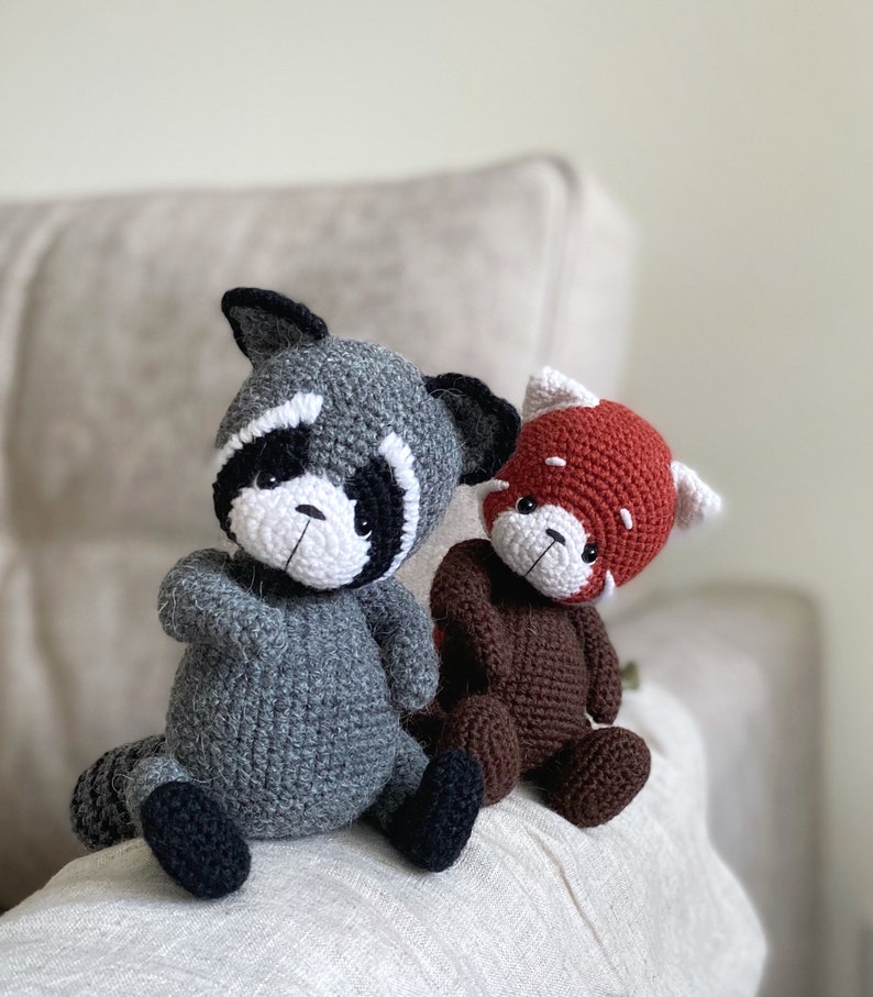 May include: Two crocheted stuffed animals, a gray raccoon and a brown red panda, sitting on a white surface.