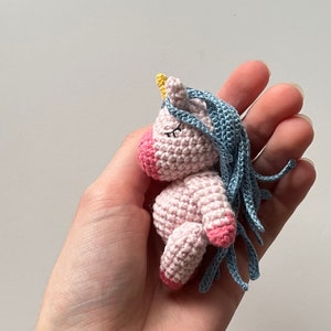 May include: A small pink crocheted unicorn with a blue mane and a yellow horn. The unicorn is lying on its side with its eyes closed.