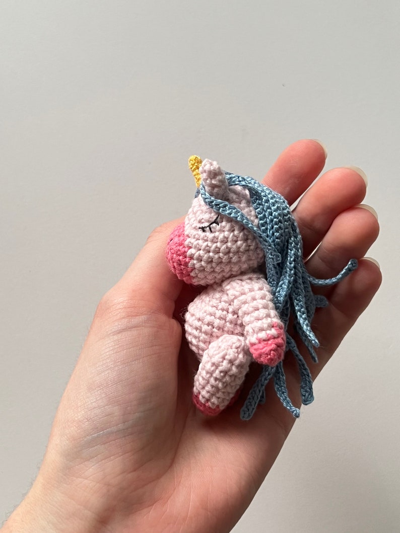May include: A small pink crocheted unicorn with a blue mane and a yellow horn. The unicorn is lying on its side with its eyes closed.