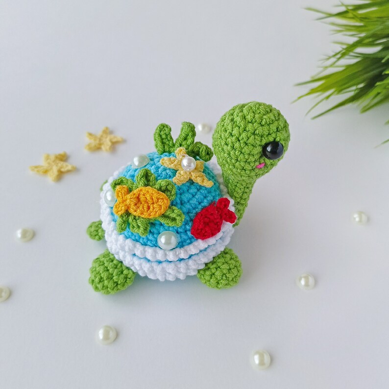 May include: A crocheted green turtle with a blue shell decorated with yellow fish, red flowers, and white pearls. The turtle has black eyes and a pink mouth.
