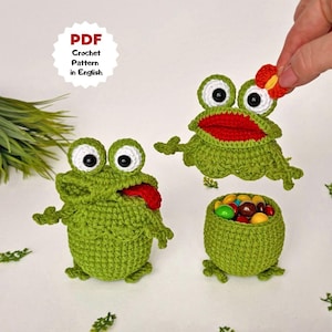 Può includere: Creazioni di rane verdi all'uncinetto. Una rana ha la lingua di fuori, un'altra ha la bocca aperta e una terza è una ciotola piena di caramelle colorate. L'immagine include il testo "PDF Crochet Pattern in English".