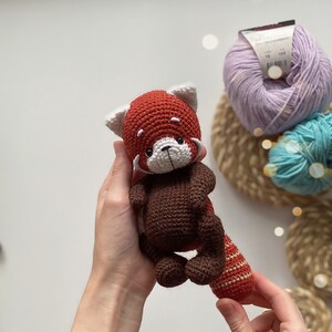May include: A crocheted red panda amigurumi toy with brown fur, white facial features, and a striped tail. The toy is being held by a person's hands.