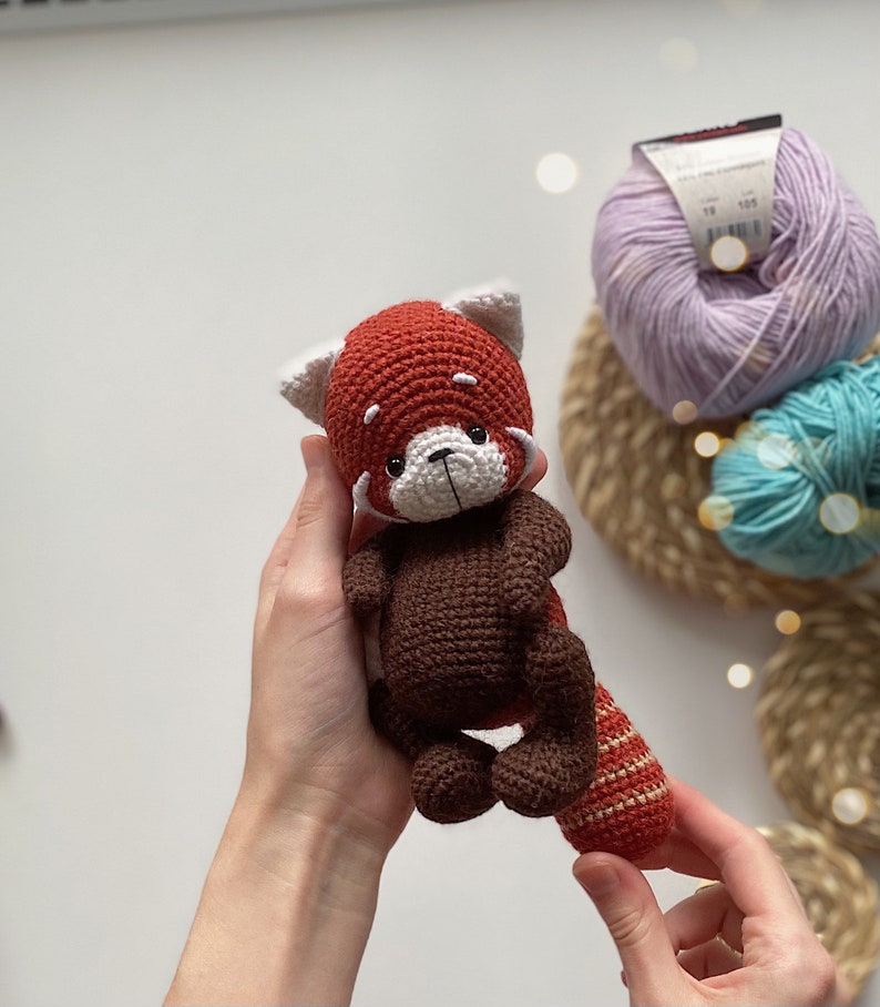 May include: A crocheted red panda amigurumi toy with brown fur, white facial features, and a striped tail. The toy is being held by a person's hands.