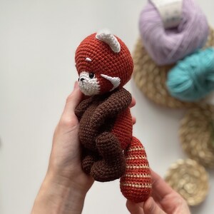 May include: A crocheted red panda plush toy with brown and white accents. The toy is being held by a person's hands.