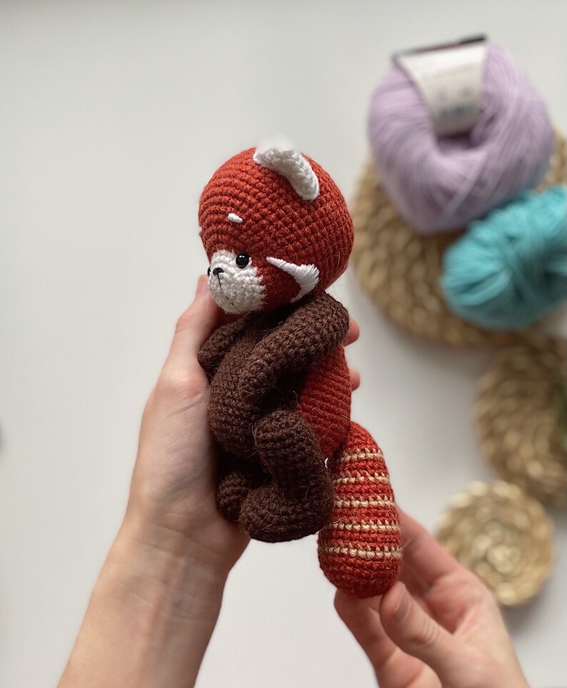May include: A crocheted red panda plush toy with brown and white accents. The toy is being held by a person's hands.