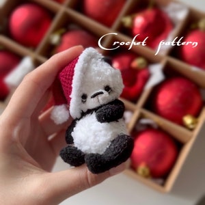May include: A small crocheted panda wearing a red Santa hat. The panda is being held in a hand and is in front of a box of red Christmas ornaments.