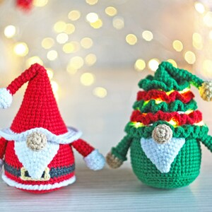 May include: Two crocheted gnomes, one in red and white and one in green and red, with white beards and light-up features. The gnomes are sitting on a wooden surface with a blurred background of white lights.